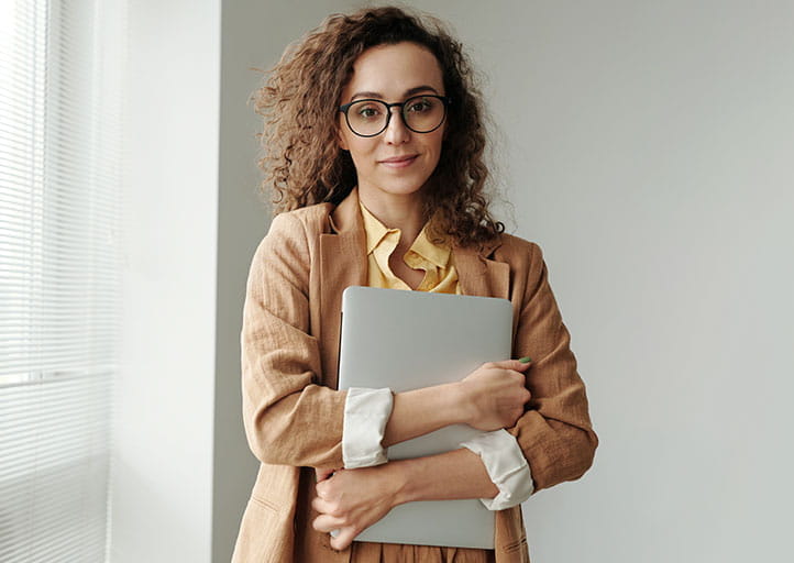 Frau mit dunklen Locken steht mit einem Laptop in der Hand.